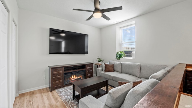 living room featuring baseboards, a glass covered fireplace, light wood-style flooring, and a ceiling fan