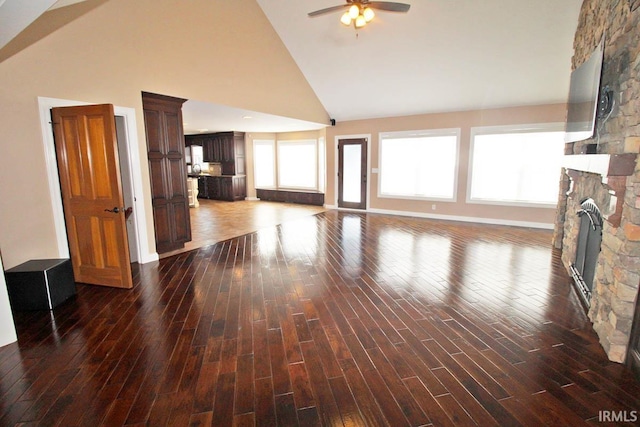 unfurnished living room with dark wood-type flooring, ceiling fan, a stone fireplace, high vaulted ceiling, and baseboards