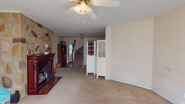 living room featuring light carpet, a fireplace, a ceiling fan, baseboards, and stairs