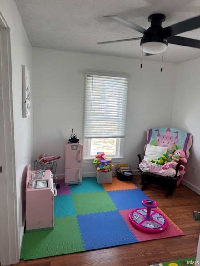 playroom with ceiling fan, a textured ceiling, baseboards, and wood finished floors