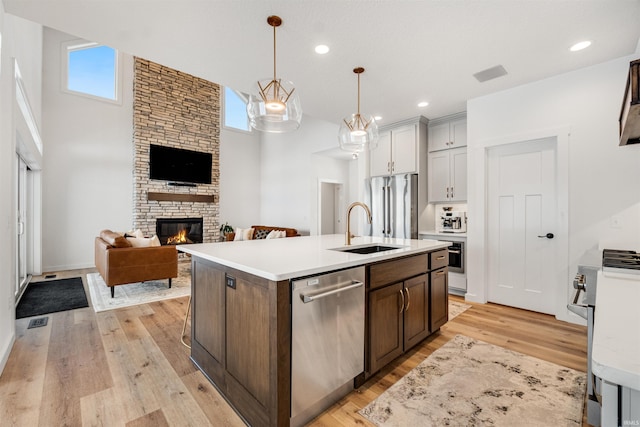 kitchen with a stone fireplace, light wood-style flooring, stainless steel appliances, an island with sink, and decorative light fixtures