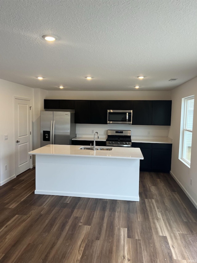 kitchen featuring a center island with sink, light countertops, appliances with stainless steel finishes, a sink, and dark cabinets