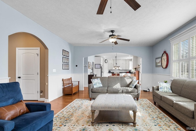 living room with arched walkways, ceiling fan, a textured ceiling, wood finished floors, and wainscoting