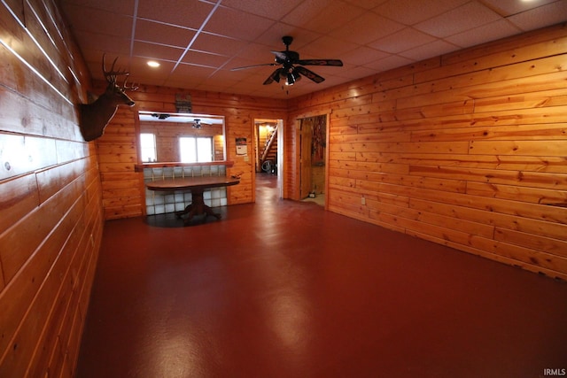 interior space with a ceiling fan, a drop ceiling, and wooden walls