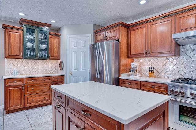 kitchen featuring brown cabinets, stainless steel appliances, glass insert cabinets, light tile patterned flooring, and under cabinet range hood