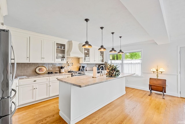 kitchen featuring glass insert cabinets, freestanding refrigerator, white cabinets, an island with sink, and premium range hood