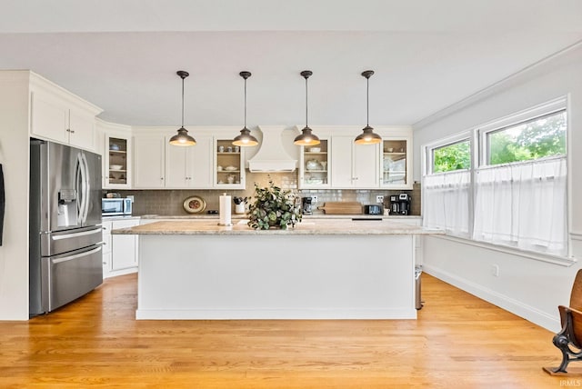 kitchen featuring pendant lighting, custom exhaust hood, stainless steel appliances, glass insert cabinets, and a kitchen island with sink