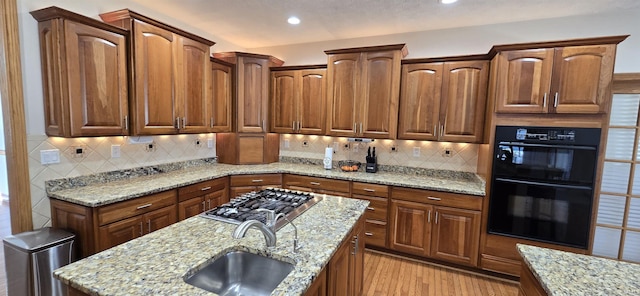 kitchen featuring dobule oven black, light wood-style flooring, light stone counters, stainless steel gas cooktop, and a sink