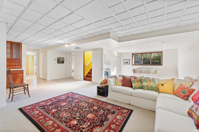 living room featuring baseboards, stairway, a drop ceiling, and light colored carpet