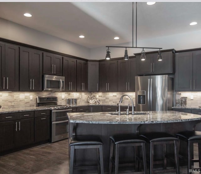 kitchen with a center island with sink, light stone counters, stainless steel appliances, and dark brown cabinets