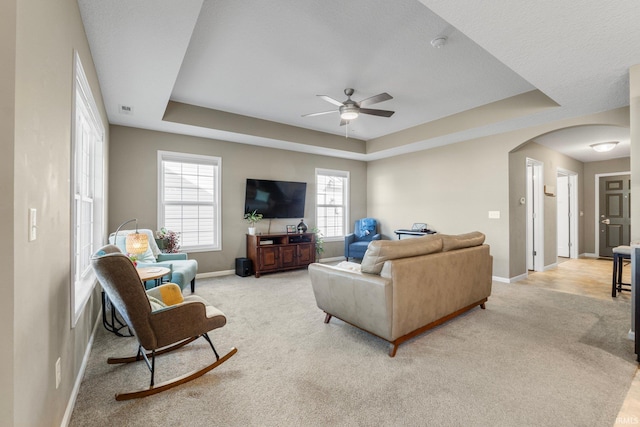 living room featuring a tray ceiling, light carpet, arched walkways, and baseboards