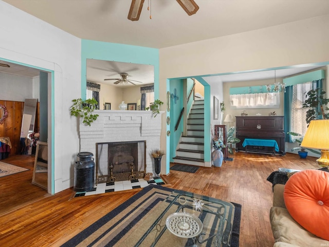 living area featuring a brick fireplace, ceiling fan, stairway, and wood finished floors