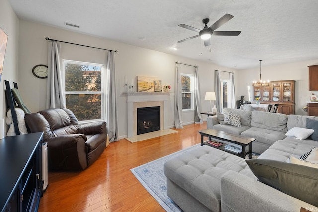 living room with visible vents, a fireplace with flush hearth, a textured ceiling, light wood-type flooring, and ceiling fan with notable chandelier