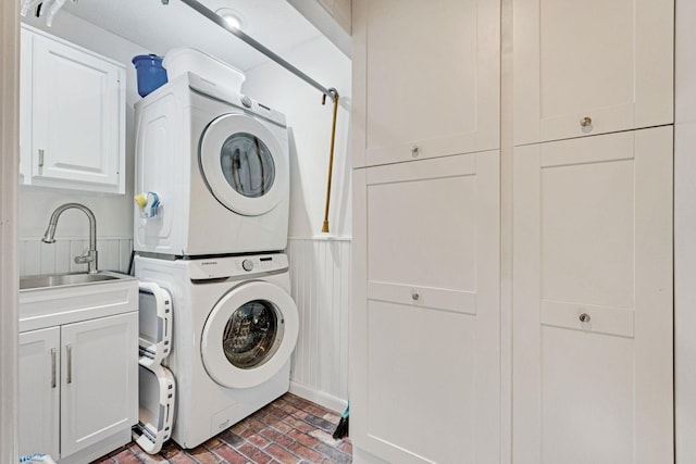 laundry area featuring brick floor, cabinet space, stacked washer / dryer, wainscoting, and a sink