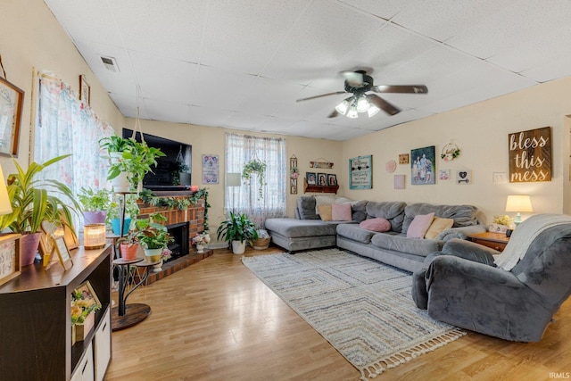 living area with a brick fireplace, ceiling fan, visible vents, and wood finished floors