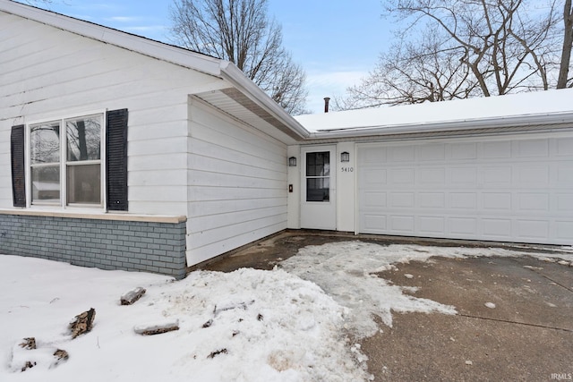 snow covered property entrance featuring driveway, brick siding, and an attached garage