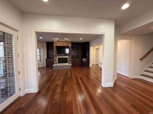 unfurnished living room featuring dark wood-style floors, recessed lighting, a brick fireplace, baseboards, and stairs