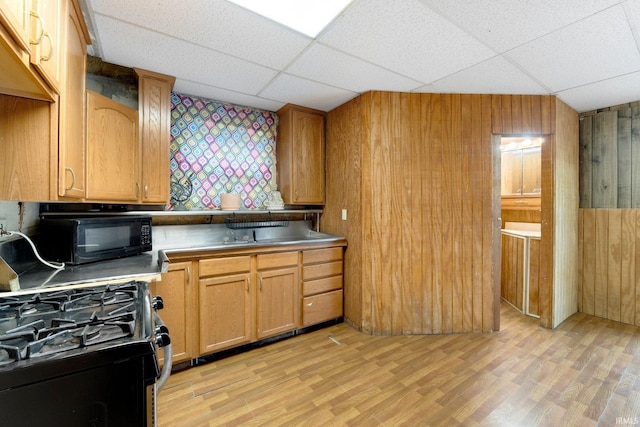 kitchen with black microwave, a drop ceiling, wooden walls, light wood finished floors, and gas range