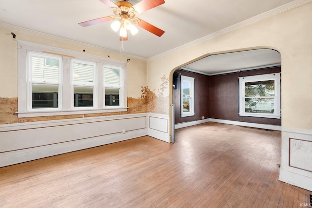 empty room with arched walkways, ceiling fan, wood finished floors, ornamental molding, and wainscoting