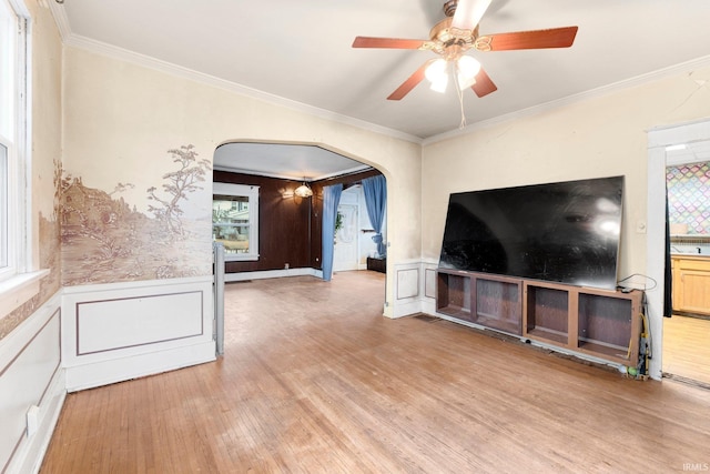 unfurnished living room with arched walkways, ceiling fan, wood finished floors, wainscoting, and crown molding