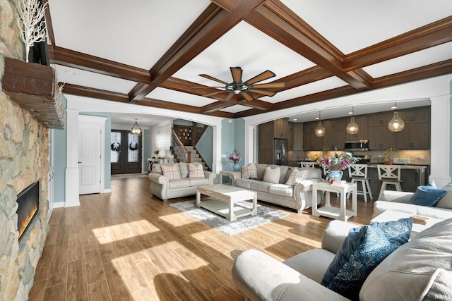 living area featuring light wood-type flooring, coffered ceiling, a stone fireplace, and stairs