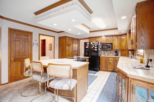 kitchen with tile counters, stainless steel microwave, black fridge with ice dispenser, brown cabinetry, and a sink