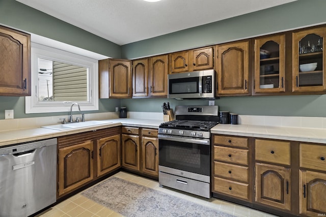 kitchen with tile countertops, stainless steel appliances, a sink, brown cabinetry, and glass insert cabinets