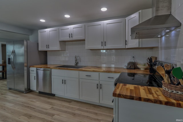 kitchen with light wood finished floors, appliances with stainless steel finishes, a sink, wood counters, and wall chimney range hood