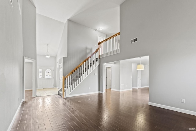 unfurnished living room featuring wood finished floors, a towering ceiling, visible vents, stairs, and baseboards