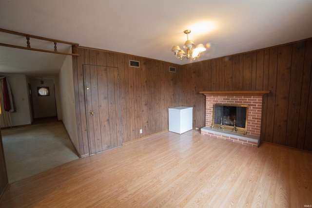 unfurnished living room featuring wooden walls, visible vents, light wood-style floors, an inviting chandelier, and a brick fireplace