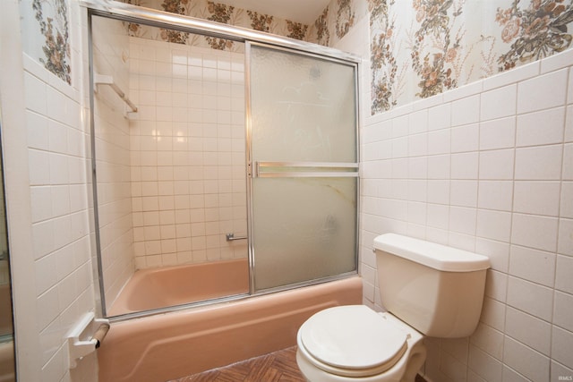 bathroom with toilet, a wainscoted wall, tile walls, combined bath / shower with glass door, and wallpapered walls