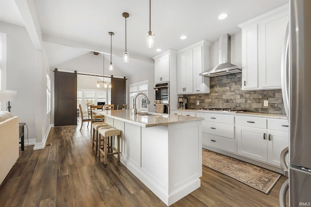 kitchen with a barn door, tasteful backsplash, dark wood finished floors, wall chimney range hood, and a sink