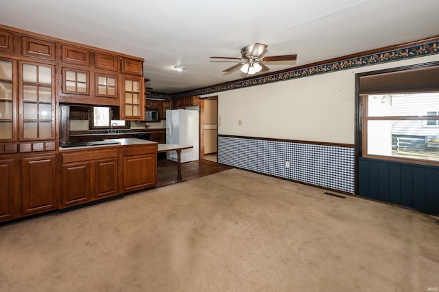 kitchen with wainscoting, ceiling fan, glass insert cabinets, brown cabinets, and white fridge with ice dispenser