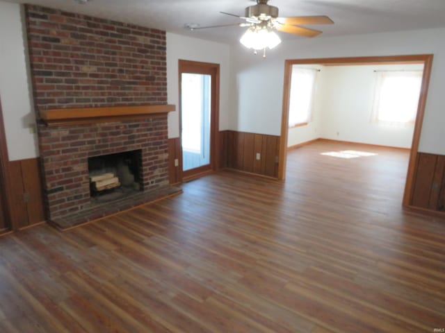 unfurnished living room featuring wainscoting, ceiling fan, wood finished floors, wood walls, and a brick fireplace