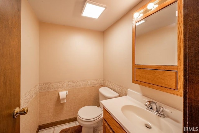 half bathroom with toilet, vanity, tile walls, wainscoting, and tile patterned floors