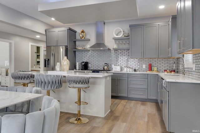 kitchen with wall chimney exhaust hood, a kitchen breakfast bar, gray cabinets, stainless steel refrigerator with ice dispenser, and open shelves