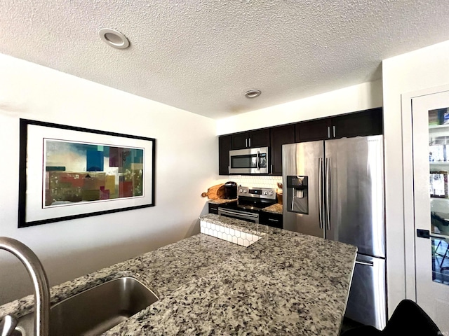 kitchen with stone counters, tasteful backsplash, appliances with stainless steel finishes, a sink, and dark cabinets