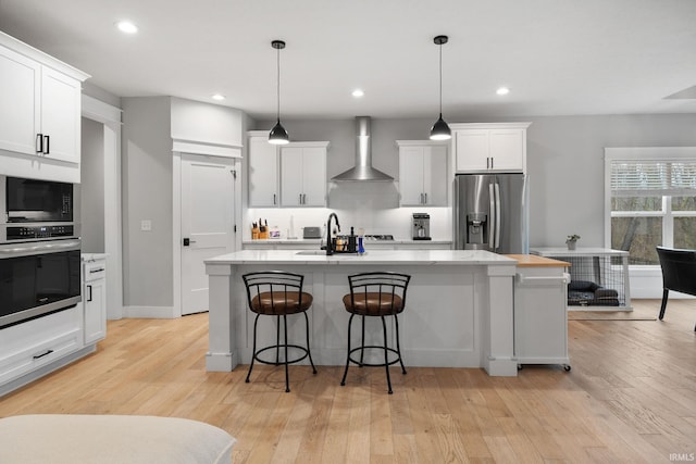 kitchen featuring a sink, white cabinetry, appliances with stainless steel finishes, wall chimney range hood, and light wood finished floors