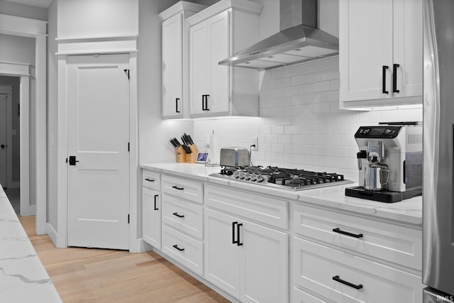 kitchen featuring light stone counters, white cabinets, appliances with stainless steel finishes, wall chimney exhaust hood, and light wood finished floors