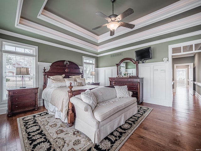 bedroom with a wainscoted wall, visible vents, ornamental molding, a tray ceiling, and wood-type flooring