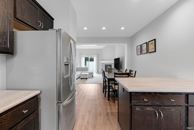 kitchen with dark brown cabinetry, stainless steel fridge, a glass covered fireplace, light wood-style flooring, and light countertops