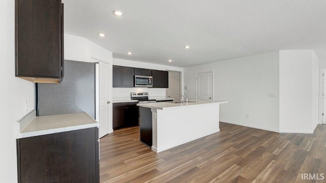 kitchen with a kitchen island with sink, stainless steel appliances, a sink, light countertops, and dark wood-style floors