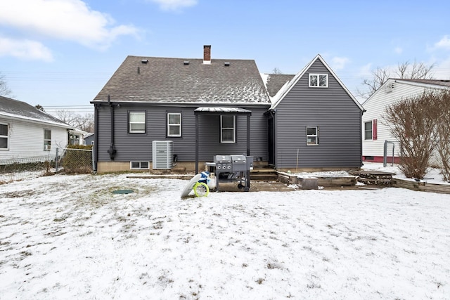 snow covered house with a chimney, fence, central AC, and roof with shingles