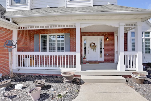 doorway to property with covered porch, brick siding, and roof with shingles