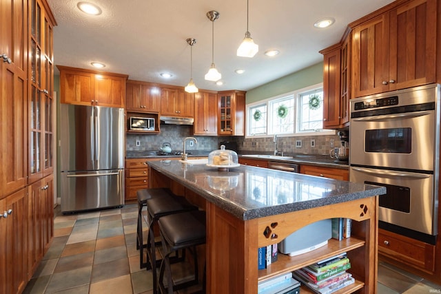 kitchen featuring under cabinet range hood, a sink, appliances with stainless steel finishes, brown cabinetry, and decorative backsplash