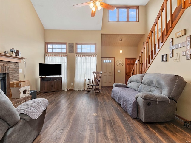 living area with dark wood-style flooring, stairway, a towering ceiling, a ceiling fan, and a brick fireplace