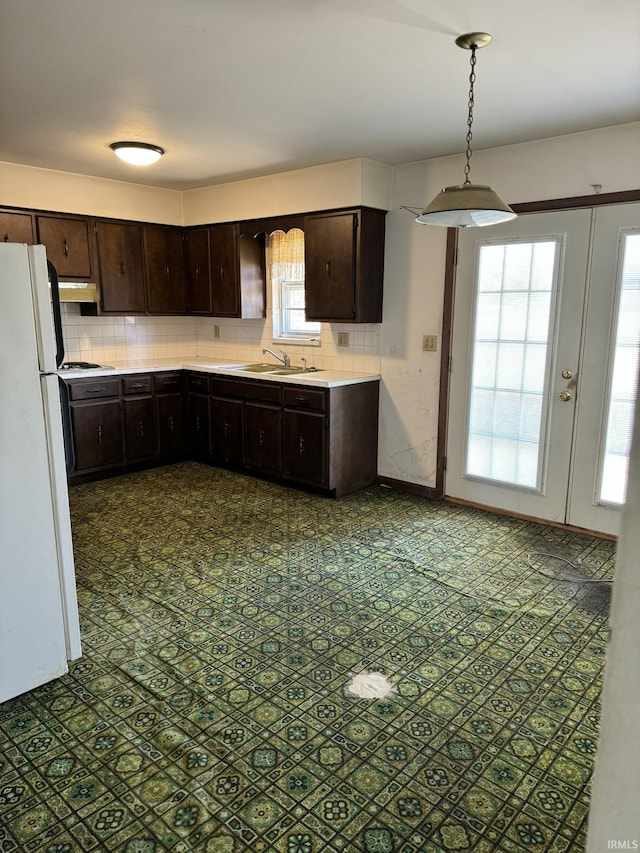 kitchen featuring dark brown cabinetry, freestanding refrigerator, light countertops, under cabinet range hood, and backsplash