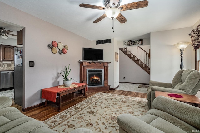 living area with visible vents, ceiling fan, wood finished floors, a warm lit fireplace, and stairs