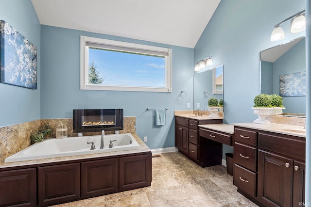 bathroom with a glass covered fireplace, a bath, two vanities, vaulted ceiling, and a sink