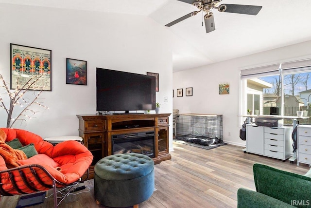 living area featuring baseboards, vaulted ceiling, wood finished floors, a glass covered fireplace, and a ceiling fan